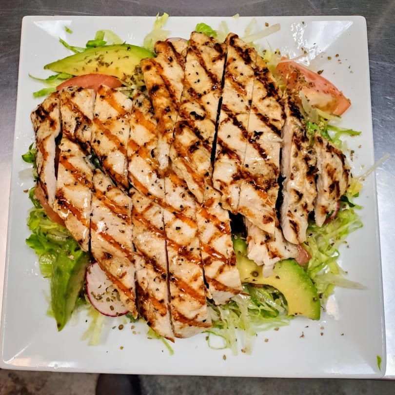 Grilled chicken salad with avocado, tomatoes, and lettuce on a white rectangular plate.