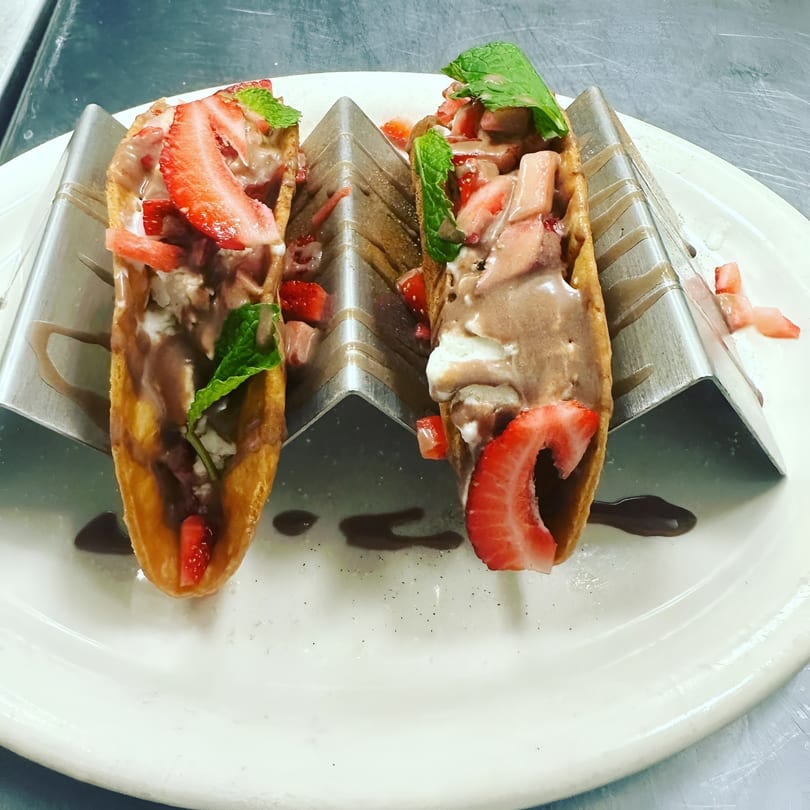 Two dessert tacos with chocolate ice cream, strawberries, and mint in metal holders on a white plate.