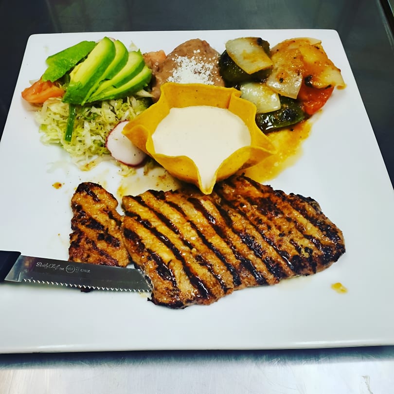 Grilled steak with avocado, salad, refried beans, and sautéed vegetables on a white plate.